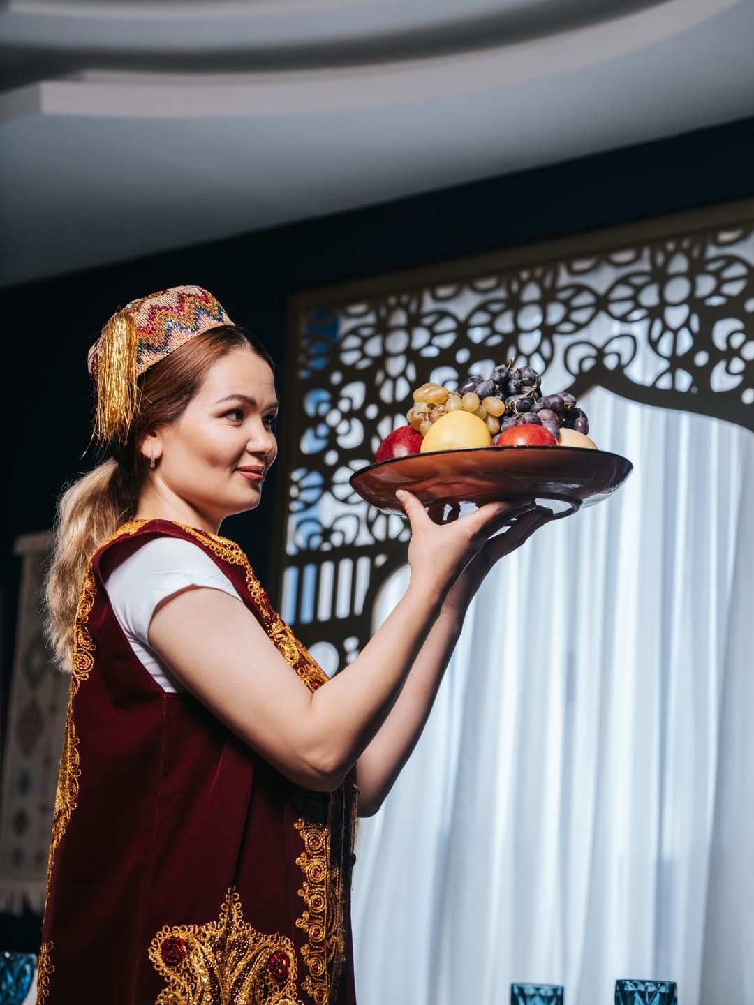 A waitress in a traditional Uzbek dress carrying a tray of fruit